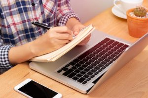 A person writing down a note on a booklet, with a laptop on the table.